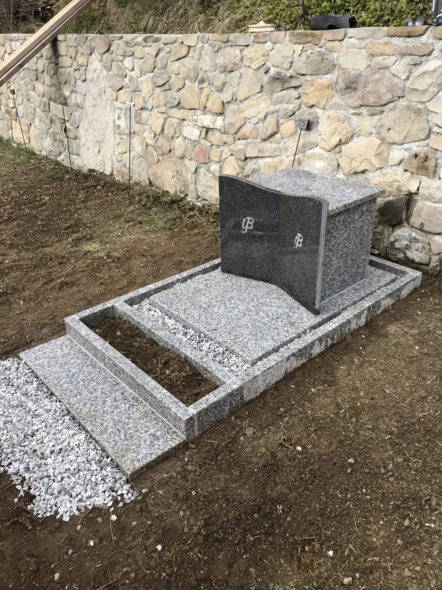 Monument funéraire en forme de livre ouvert en granit gris devant un mur de pierre.