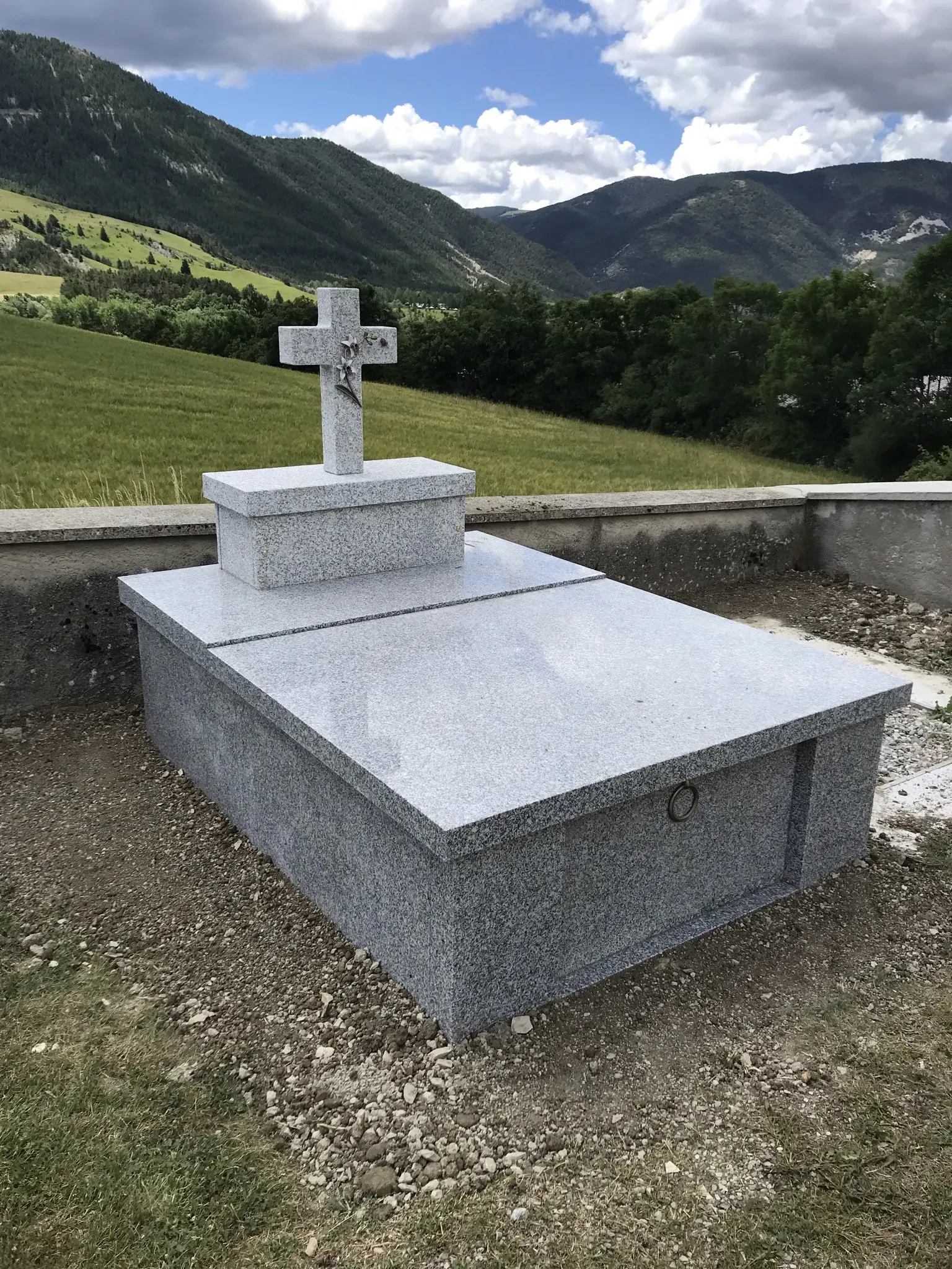Tombe moderne en granit gris avec une croix, située dans un cimetière de montagne.